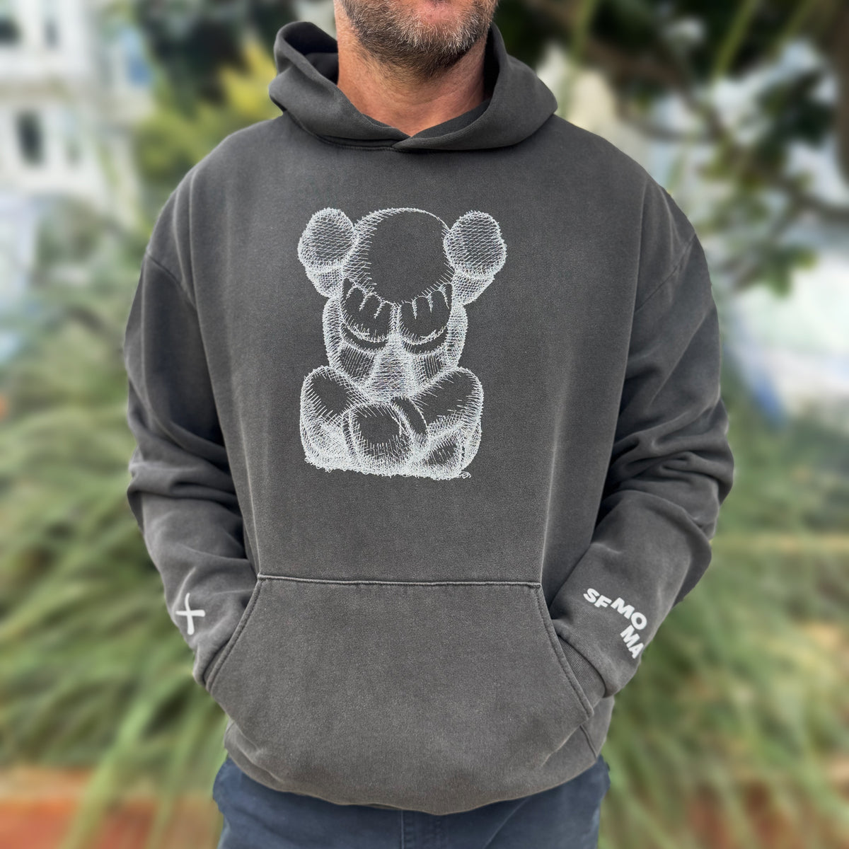 Person wearing a dark gray hoodie with a white KAWS graphic and text, standing outdoors.