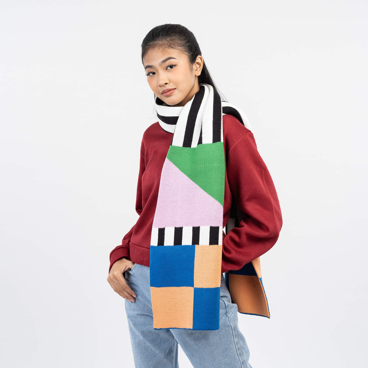 A multicolor knit scarf featuring a patchwork design with green, blue, peach, pink, and black-and-white striped sections, displayed on a light gray background.