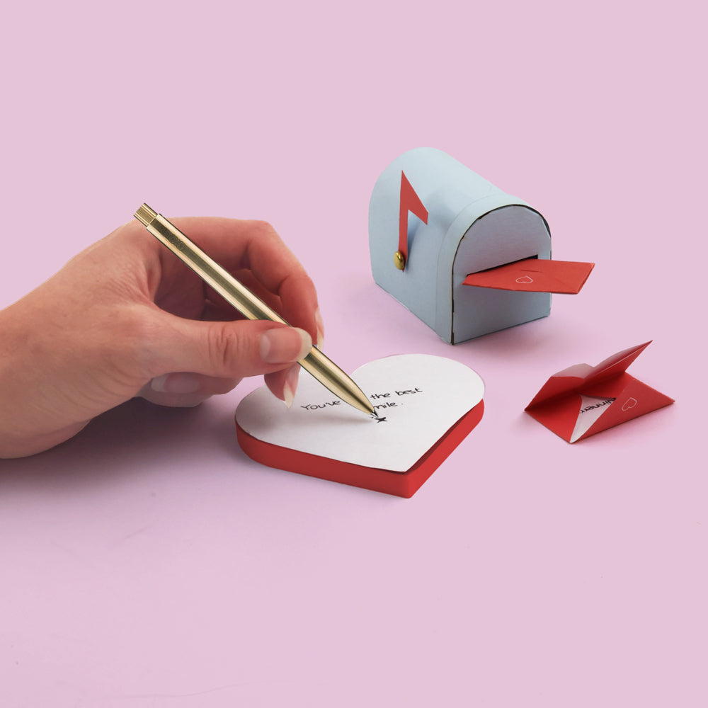 Heart-shaped notepad with a pen, small mailbox, and envelope on a pink background