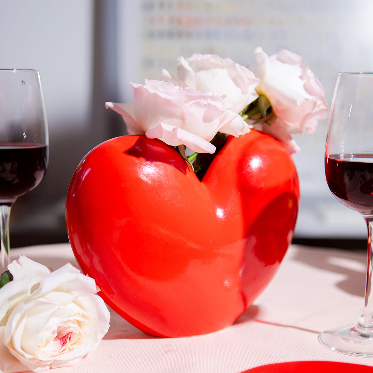 Red heart shaped vase with roses on dining table.