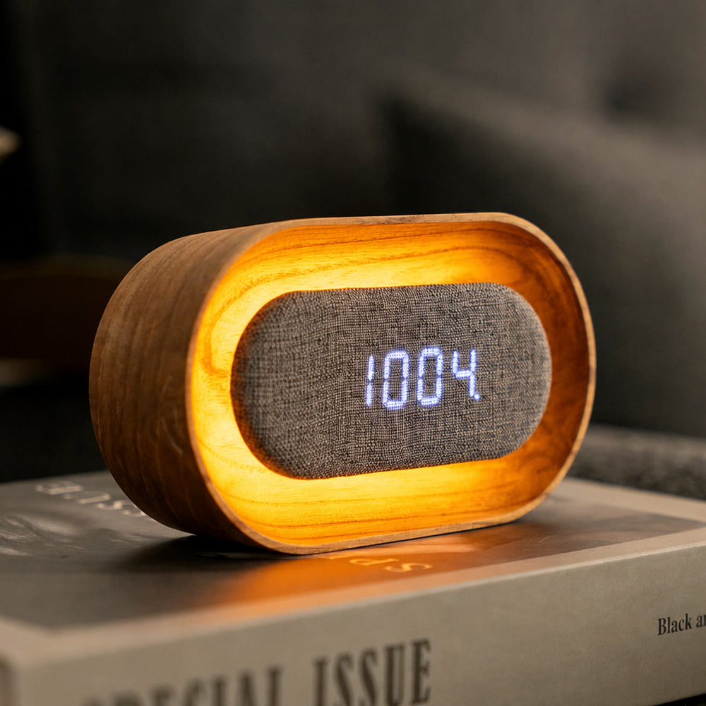A modern oval digital clock with a warm glowing wood-grain frame and a gray fabric face displaying the time 10:04 in blue LED digits, resting on a book in a cozy indoor setting.