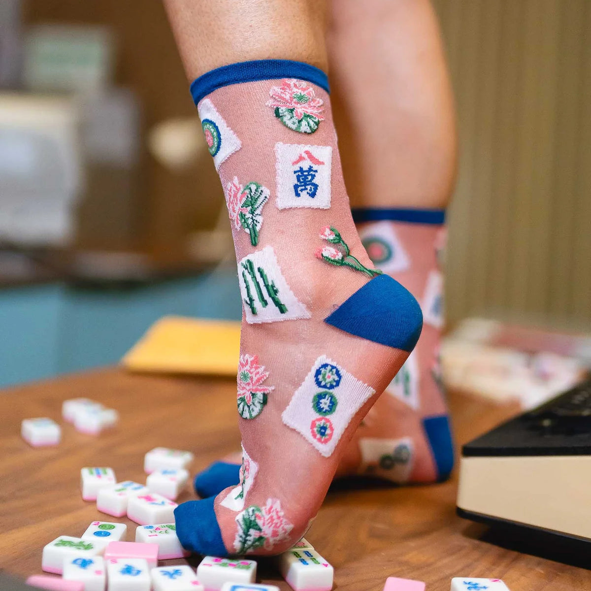 A person's legs wearing sheer pink novelty socks with embroidered mahjong tile designs and blue toe/heel caps, standing on a wooden table with scattered matching tile pieces.