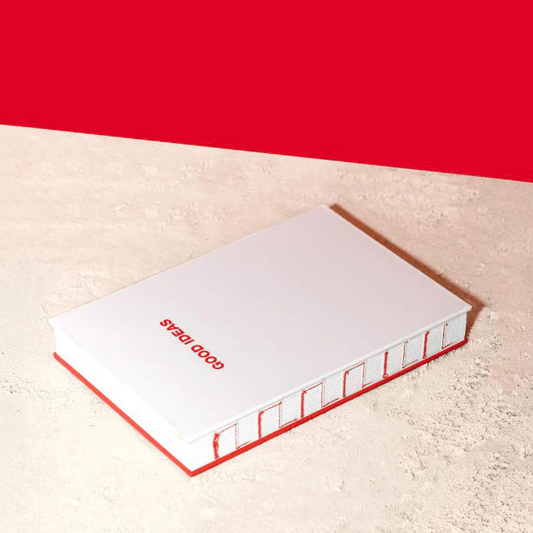 A white notebook with a red cover and red spine, lying on a textured beige surface with a solid red background. The cover features the text 'GOOD IDEAS' in red.