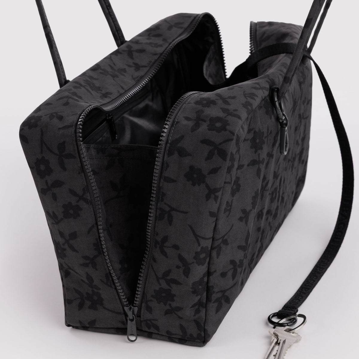 A black floral patterned bag with an open top zipper, shown on a white background with its shoulder strap and attached key visible.