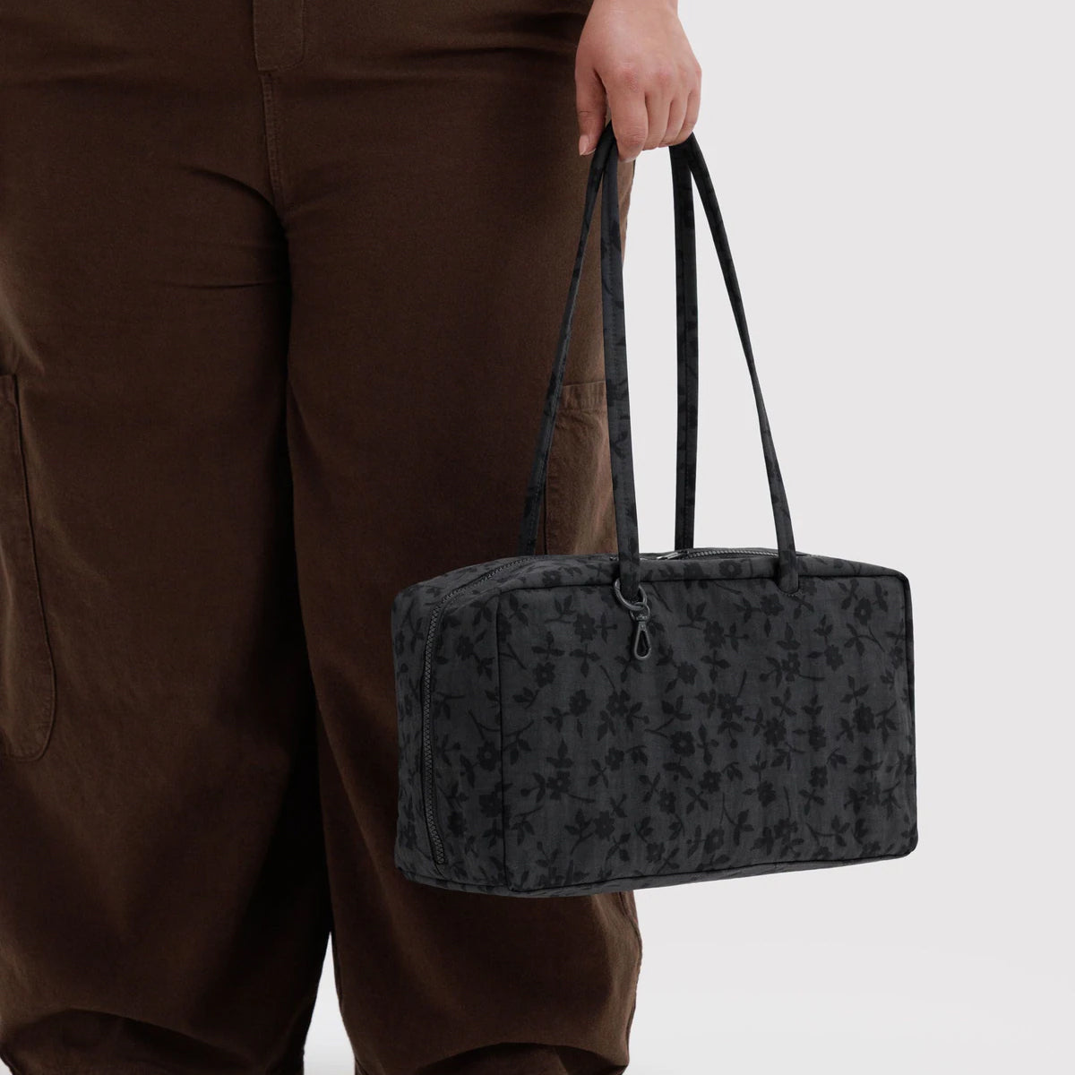 A person wearing brown pants holds a dark gray floral-patterned tote bag with black handles against a light gray background.