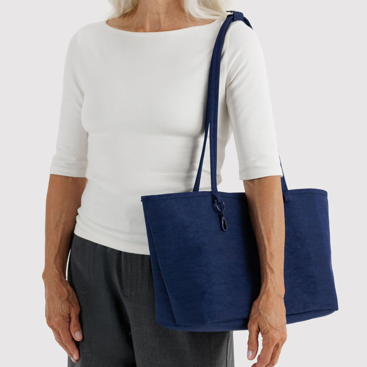 A navy blue nylon tote bag with a matching shoulder strap, carried on the shoulder of a model wearing a white 3/4-sleeve top and dark gray trousers, against a light gray studio background.