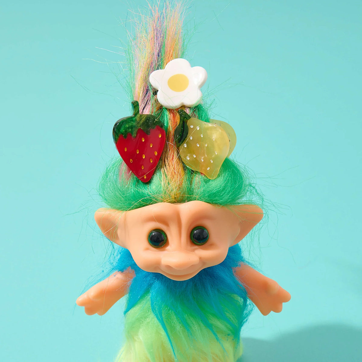 Toy figure with green hair styled with fruit and flower claiws on a light blue background