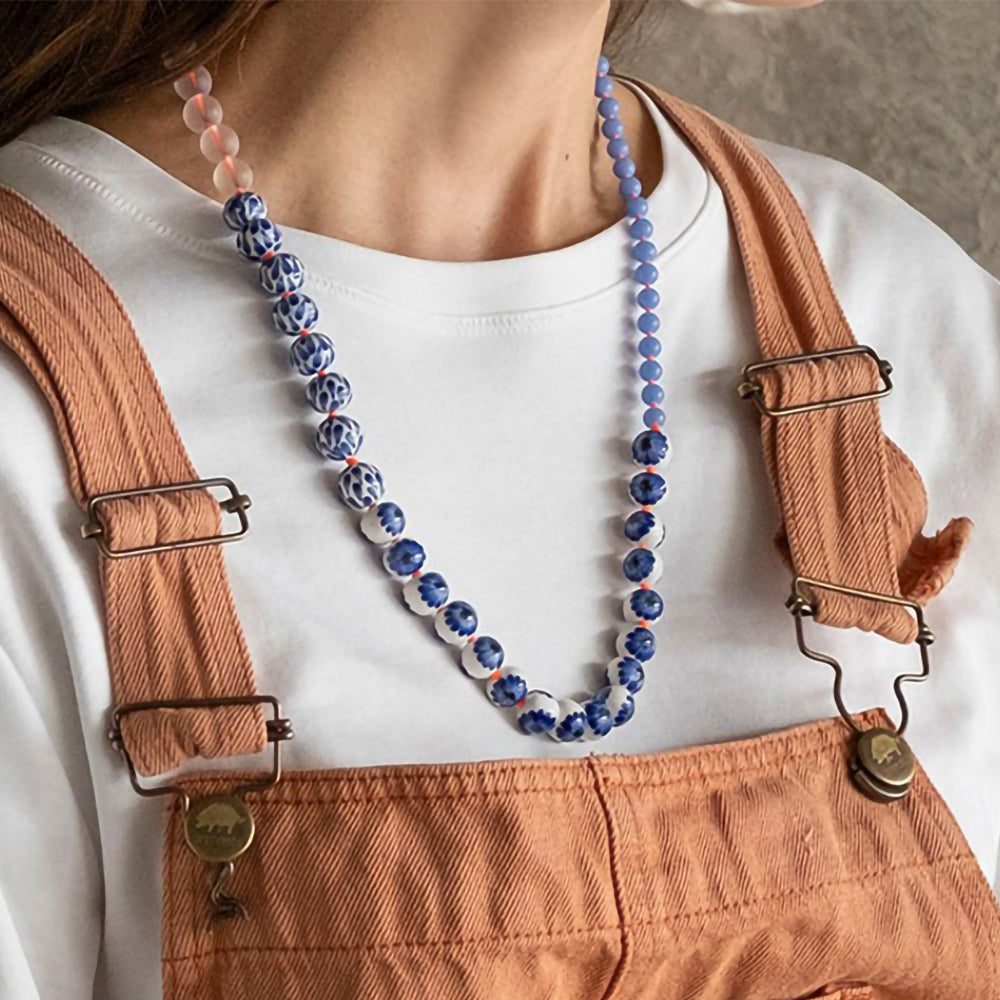 Person wearing brown overalls with a white shirt and blue beaded necklace.