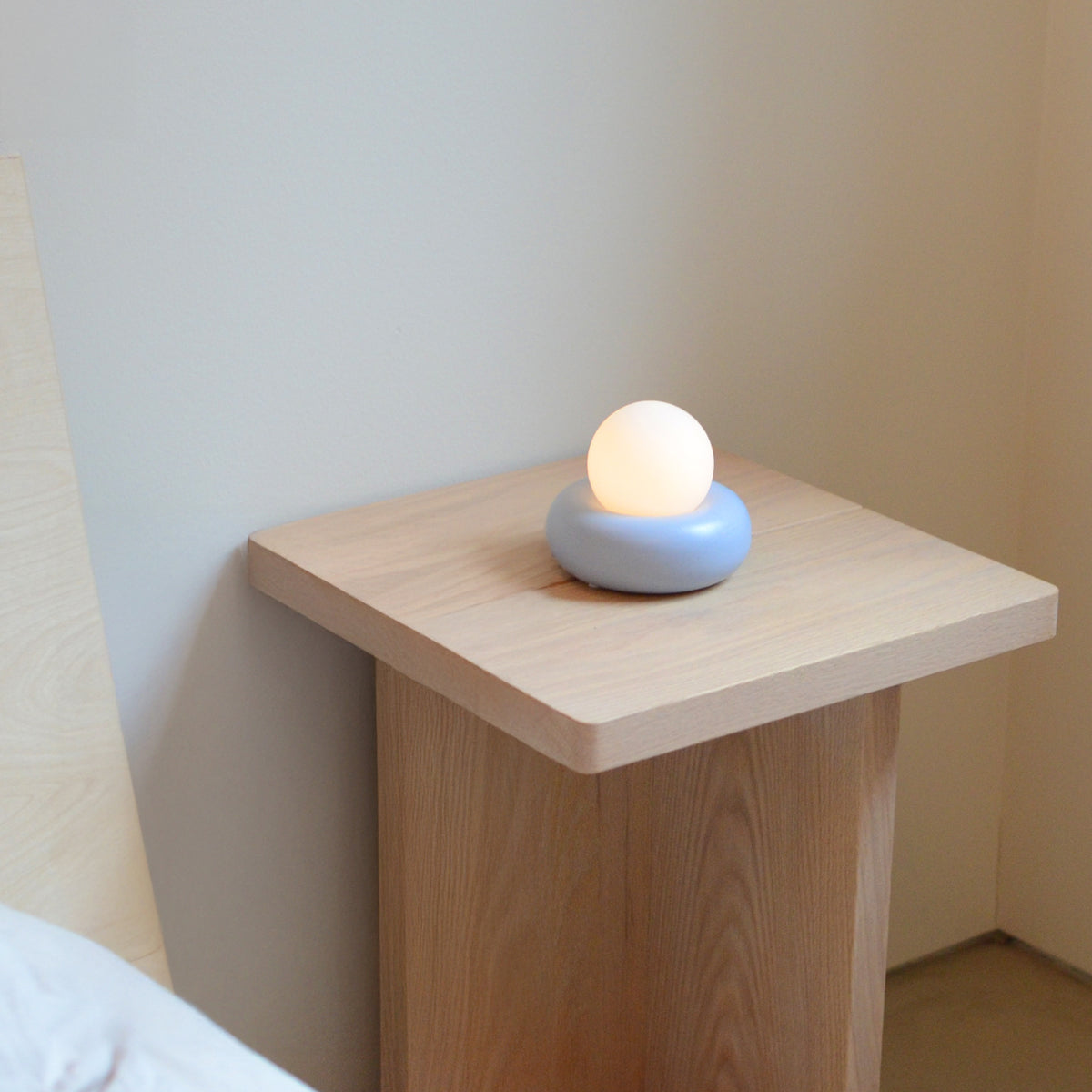 A small modern lamp with a glowing white spherical bulb resting on a light blue circular base, placed on a light wood side table in a minimalist room.