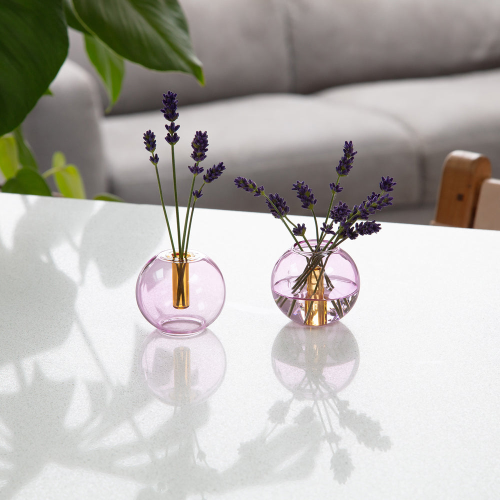 Two round pink glass vases with gold accents, each holding lavender stems, placed on a white table in a softly blurred living room setting.