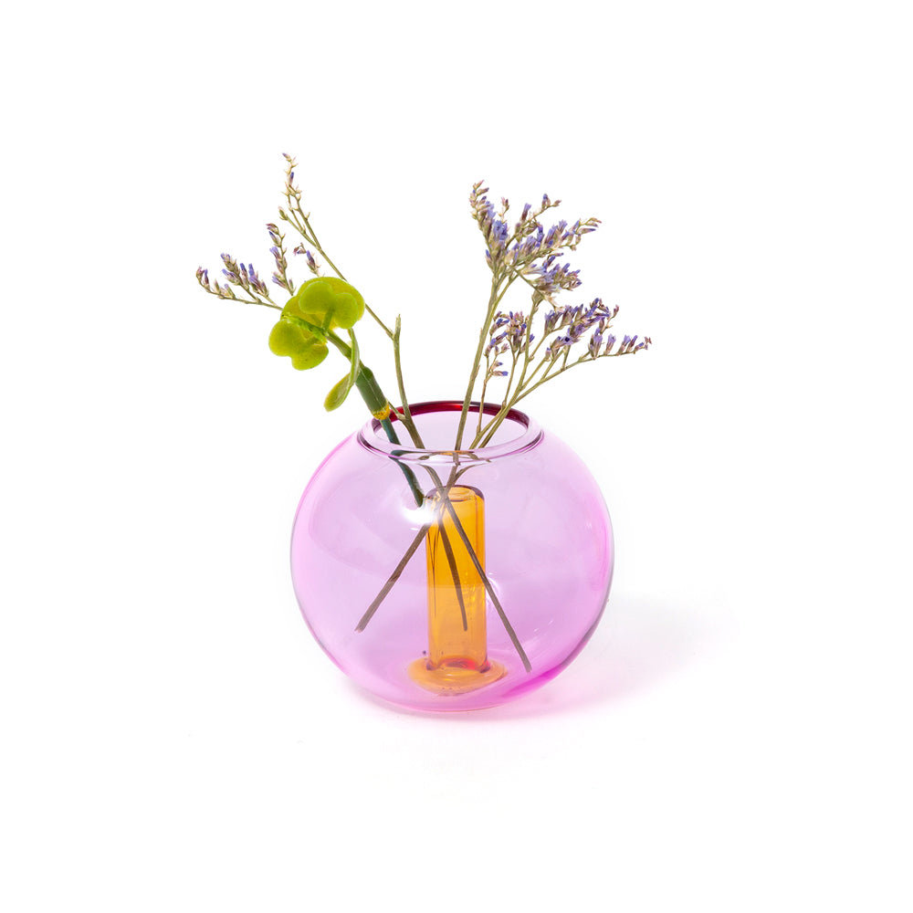 A translucent pink glass vase with a wide rounded body and a narrow neck, holding dried purple stems and a green leafy sprig against a clean white background.