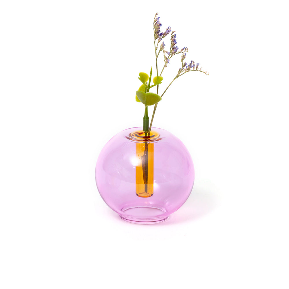 A translucent pink glass vase with a wide spherical body and a narrow central opening, holding a green stem with small purple flowers, on a clean white background.