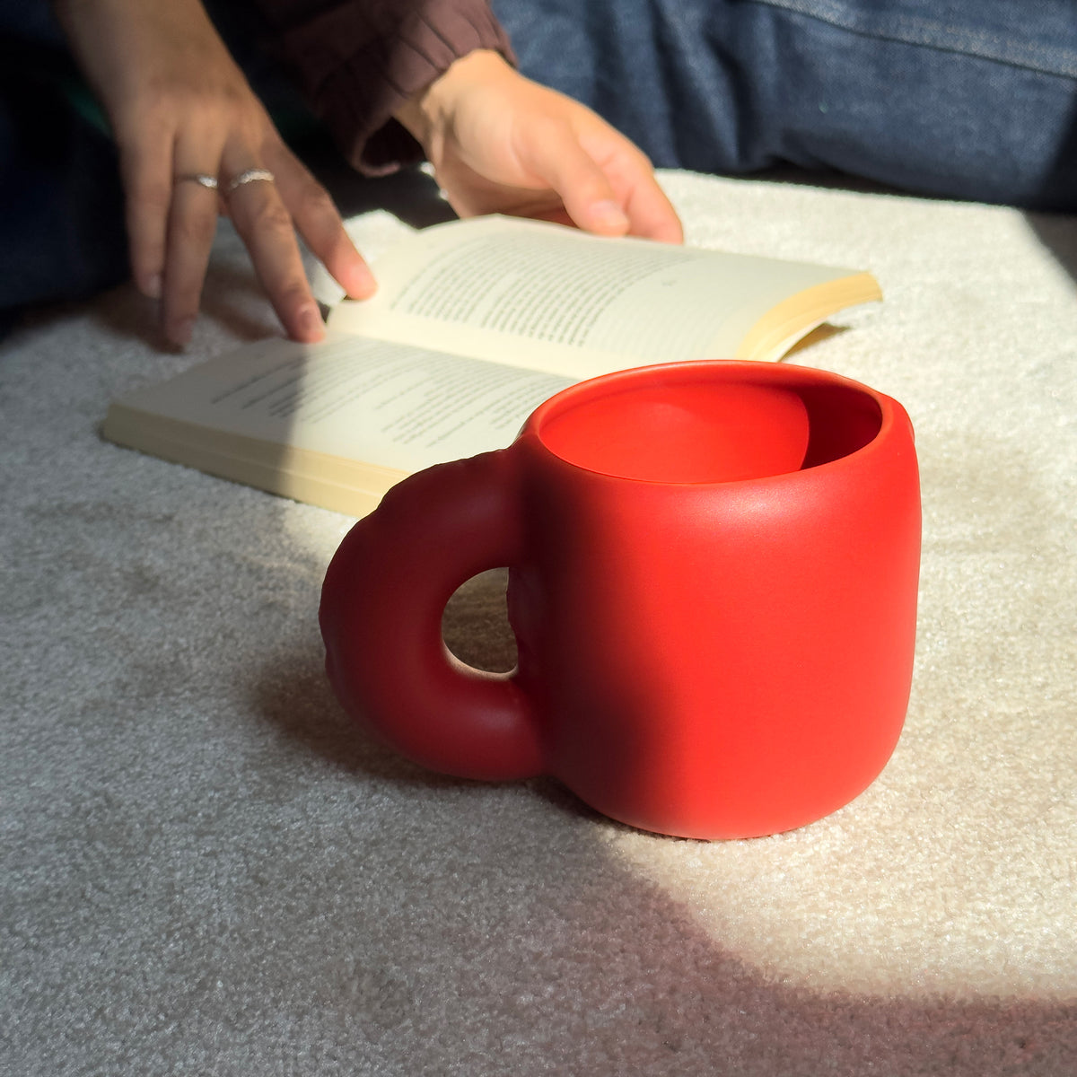 A bright red novelty mug sits on a light gray carpet next to an open book, with a person's hands and legs visible in the background.