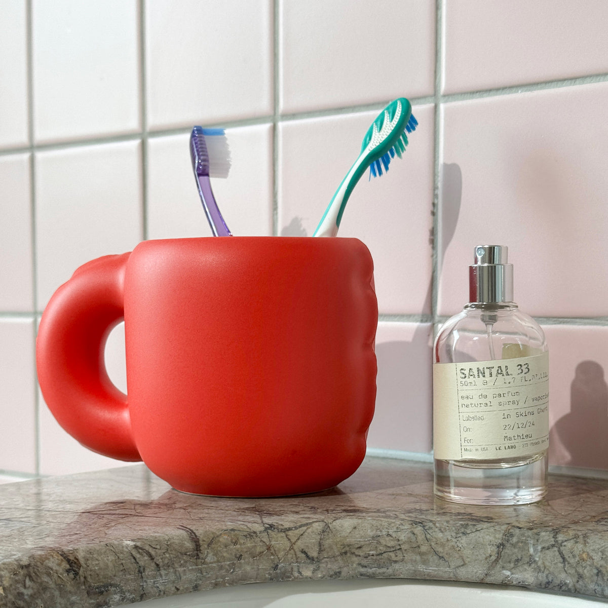 A bright red ceramic toothbrush holder shaped like a handleless mug sits on a bathroom counter next to a Santal 33 perfume bottle, with two toothbrushes inside. The background features light pink square tiles.