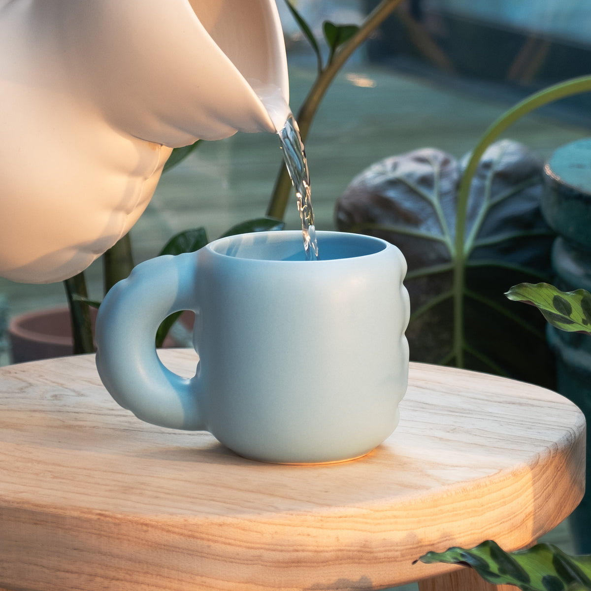 A light blue ceramic mug with a unique rounded handle sits on a wooden table, with hot water being poured into it from a white pitcher. The background features a blurred green garden setting with plants.