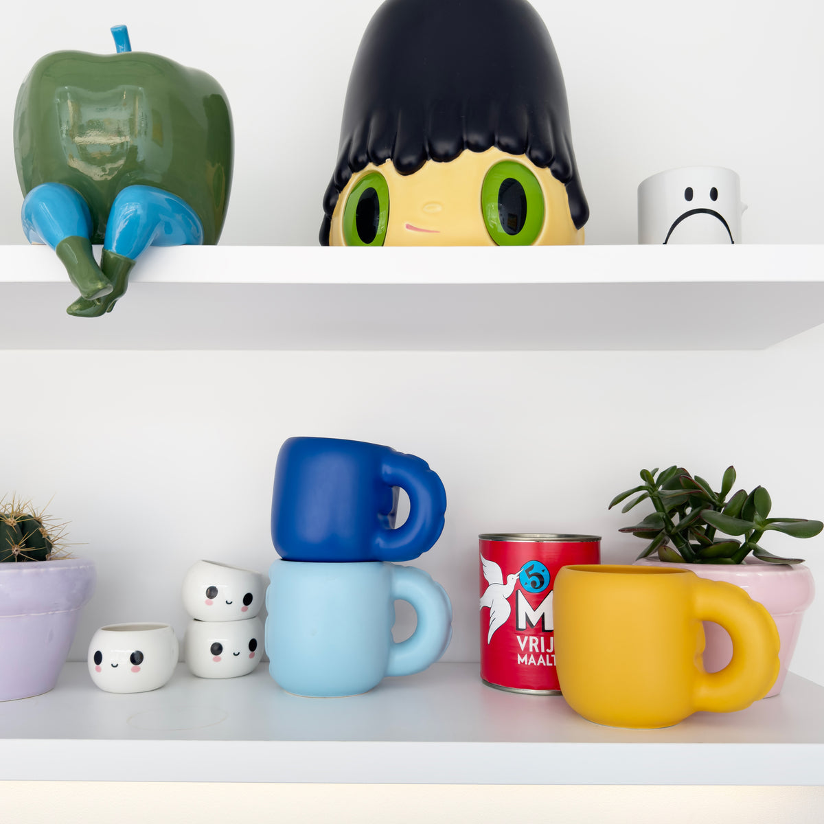 A set of cute, cartoon-style ceramic mugs displayed on white shelves. The collection includes a stack of three mugs in dark blue, light blue, and a solid yellow mug, alongside a red can-shaped mug with white lettering.