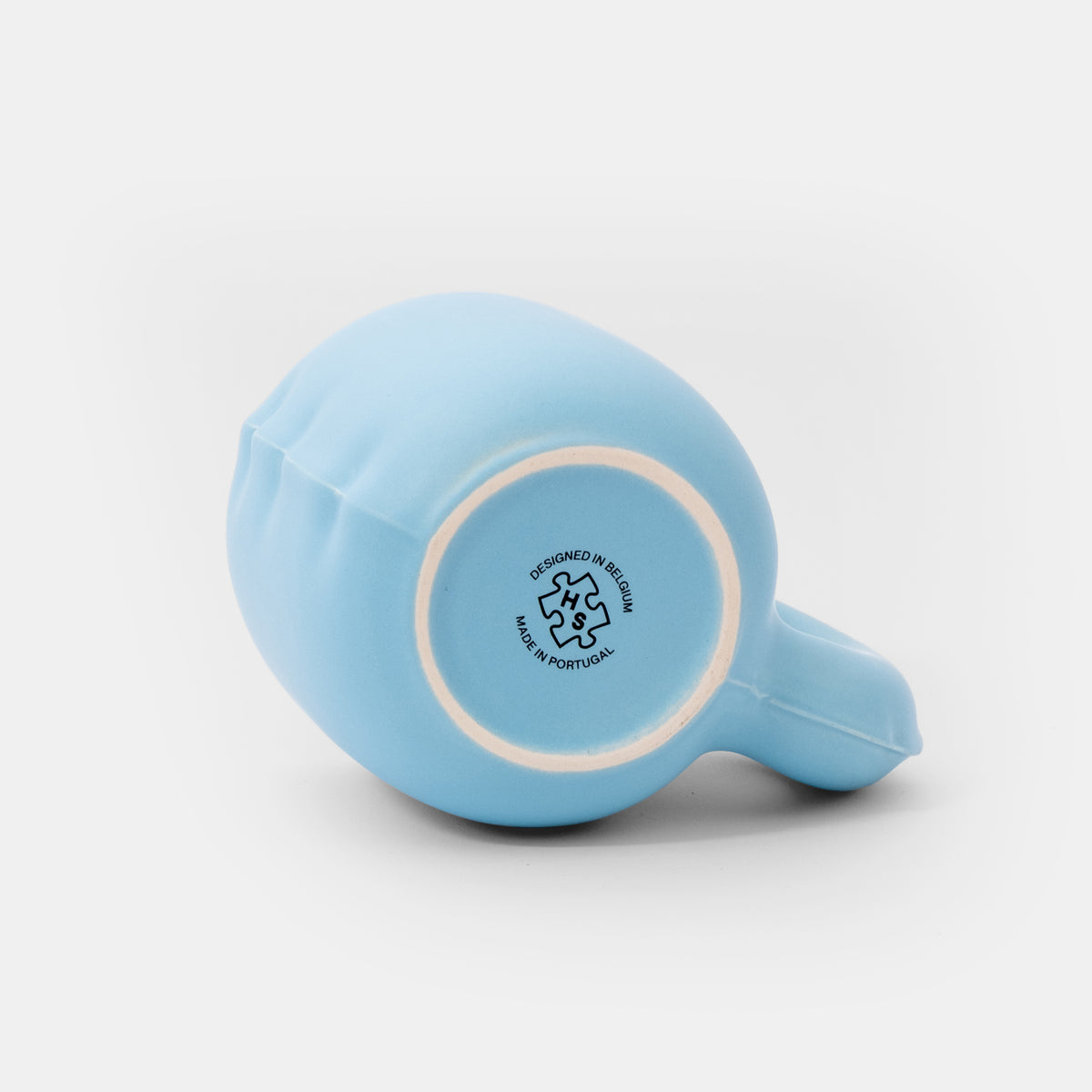 A light blue ceramic teacup turned upside down, showing its white interior and a circular black printed recycling symbol with 'DESIGNED IN BELGIUM' and 'MADE IN THE USA' on the bottom. The teacup is photographed on a clean, light gray-white background.