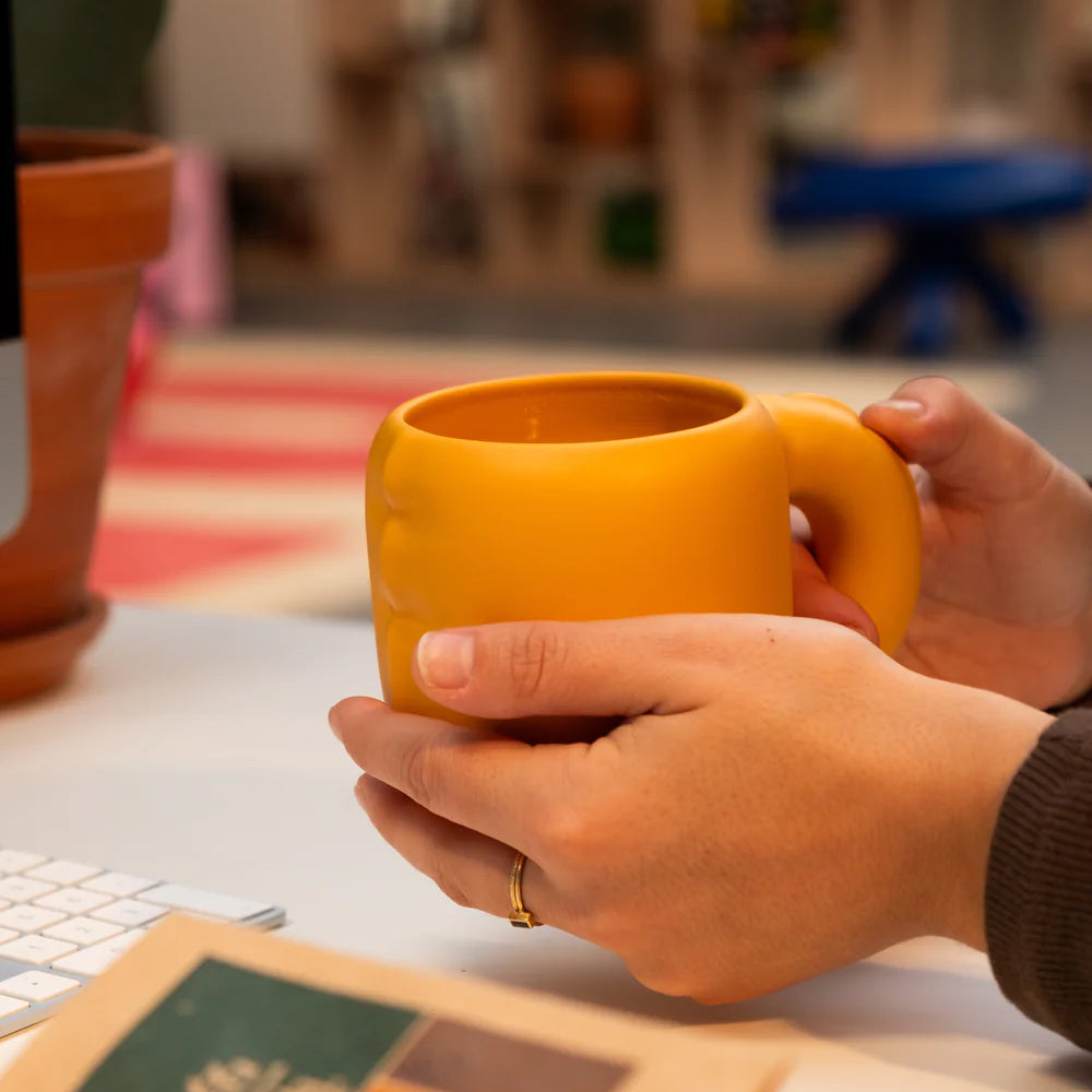 A person holding a matte mustard yellow ceramic mug with both hands at a desk, with a blurred indoor background.