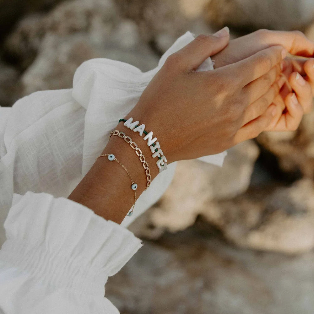A close-up of a person's wrist wearing multiple delicate bracelets, including a silver chain, a gold chain, and a thin chain with small blue evil eye charms, against a soft-focus rocky background.