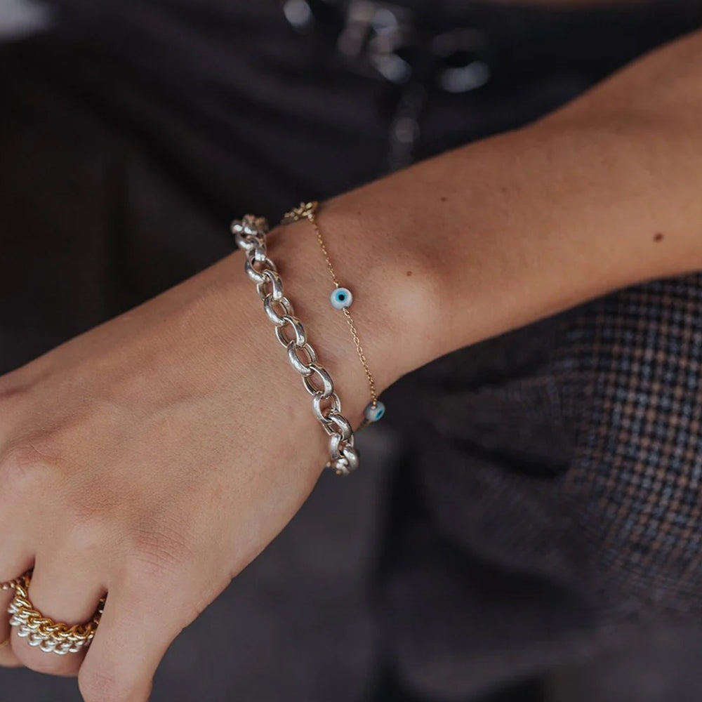 A close-up of a person's wrist wearing two delicate bracelets: a thicker silver chain bracelet and a thinner gold chain bracelet with small blue evil eye charms, shown against a dark clothing background.