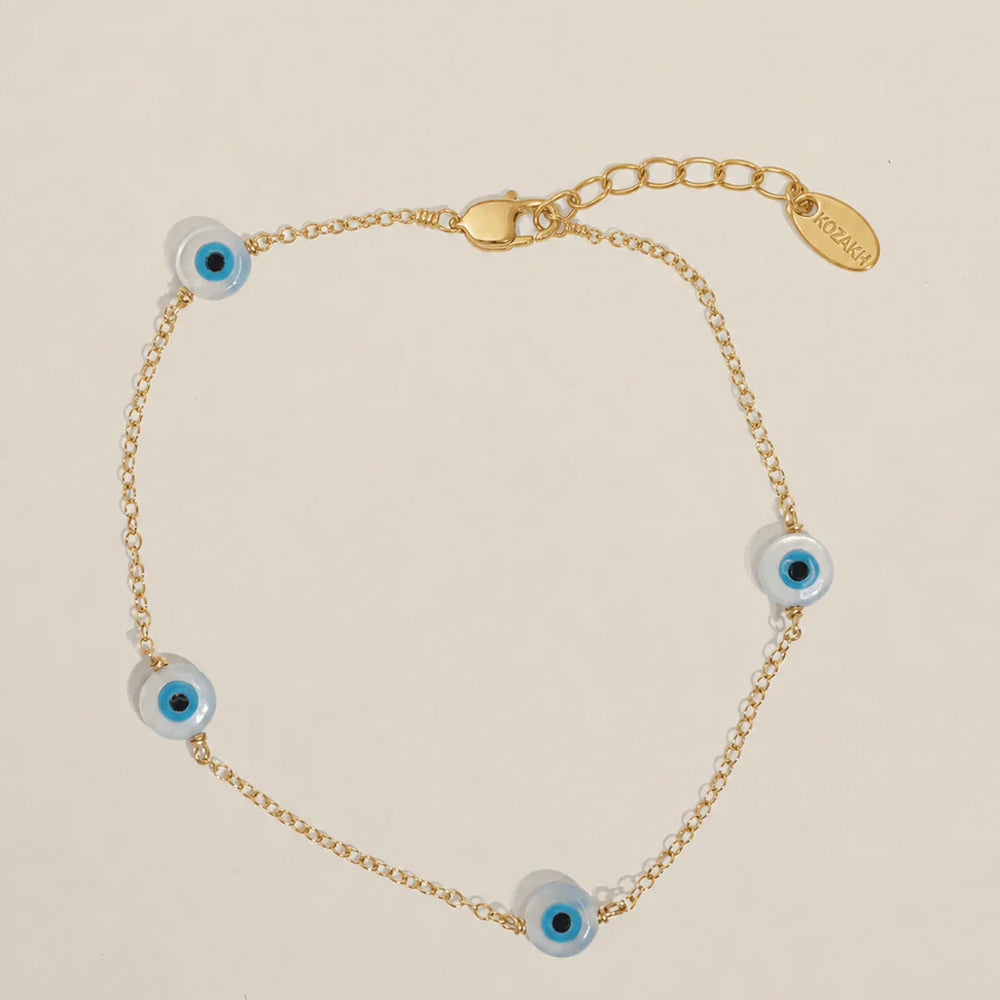 A gold chain bracelet featuring four blue and white glass evil eye beads spaced evenly along the chain, displayed on a light beige background. The chain has an adjustable lobster clasp and a gold-tone brand tag engraved.