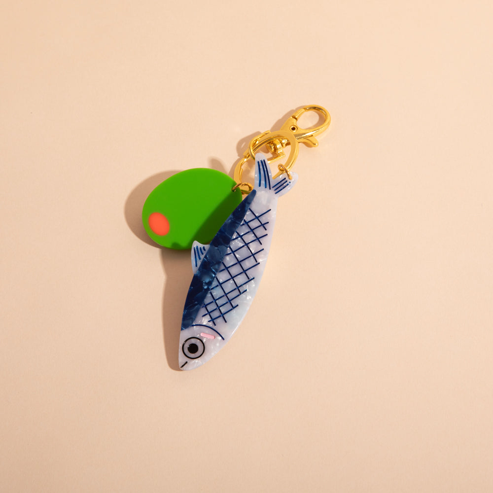 Keychain with green olive, blue anchovy, and pink label on beige background