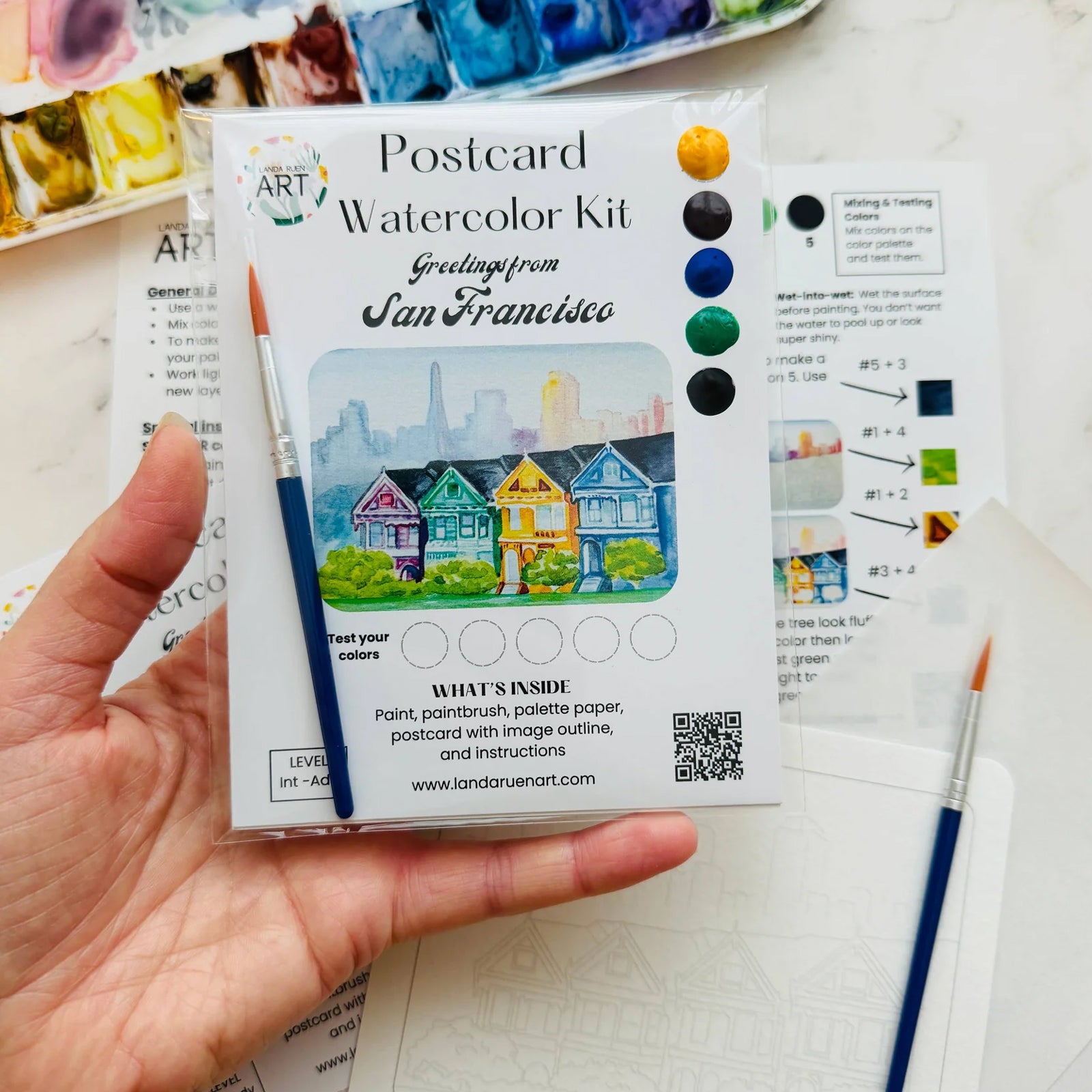 A hand holds a Landa Ruen postcard watercolor kit package, with paintbrushes and paint palettes visible on a white surface in the background.