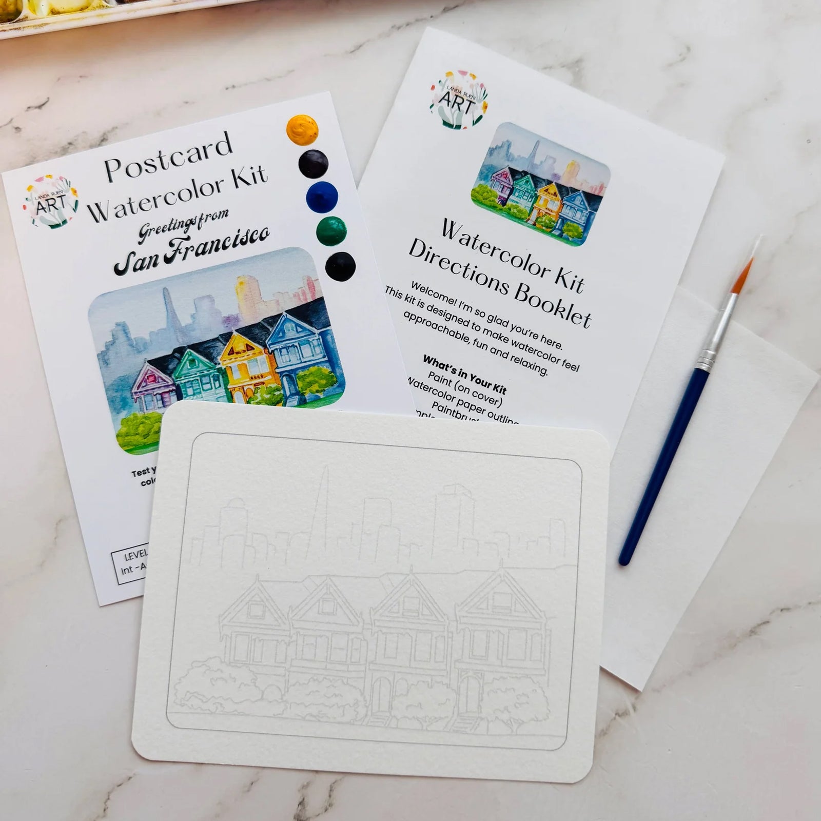 A hand holds a Landa Ruen postcard watercolor kit package, with paintbrushes and paint palettes visible on a white surface in the background.