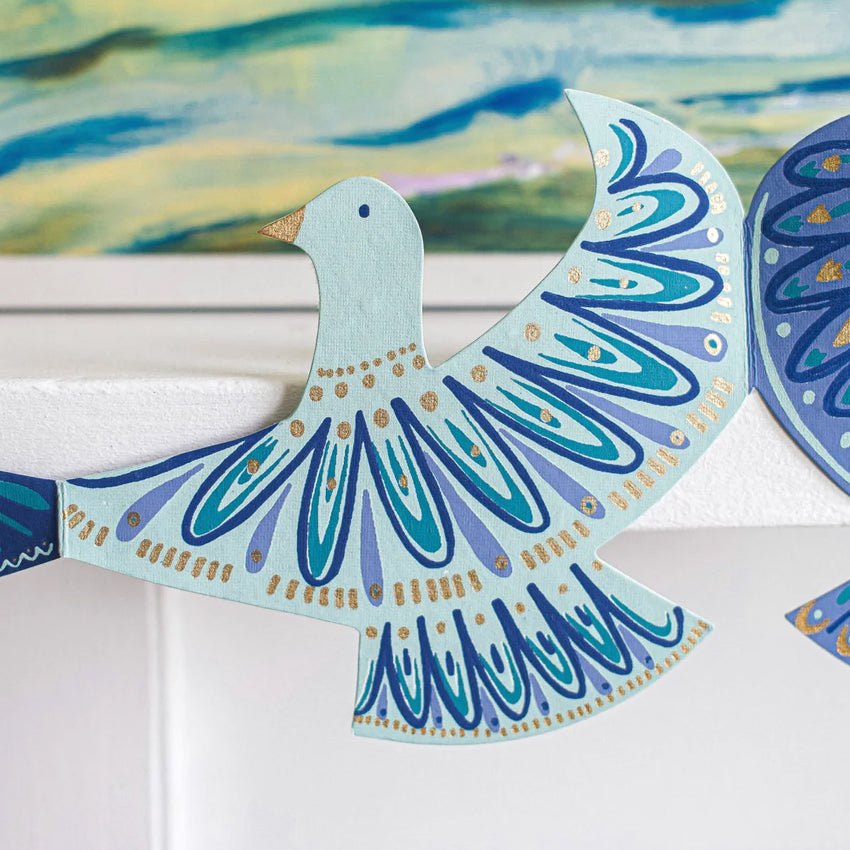 A decorative bird wall hanging or ornament with a light blue body and wings, decorated with layered blue and teal patterns and small gold accents, shown against a white wall with a colorful painting in the background.