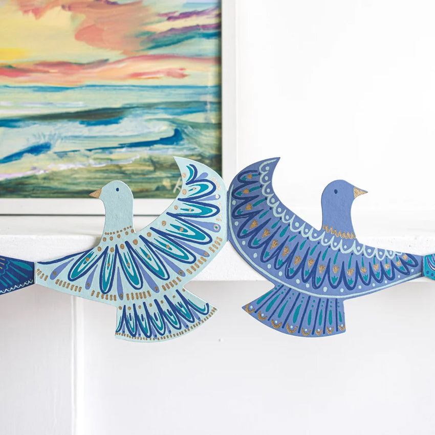 Decorative bird-shaped wall hanging garland in shades of blue, teal, and gold, shown hanging on a white wall next to a colorful framed painting.