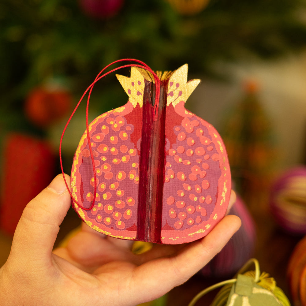Hand holding a decorative pomegranate ornament with a blurred Christmas tree background