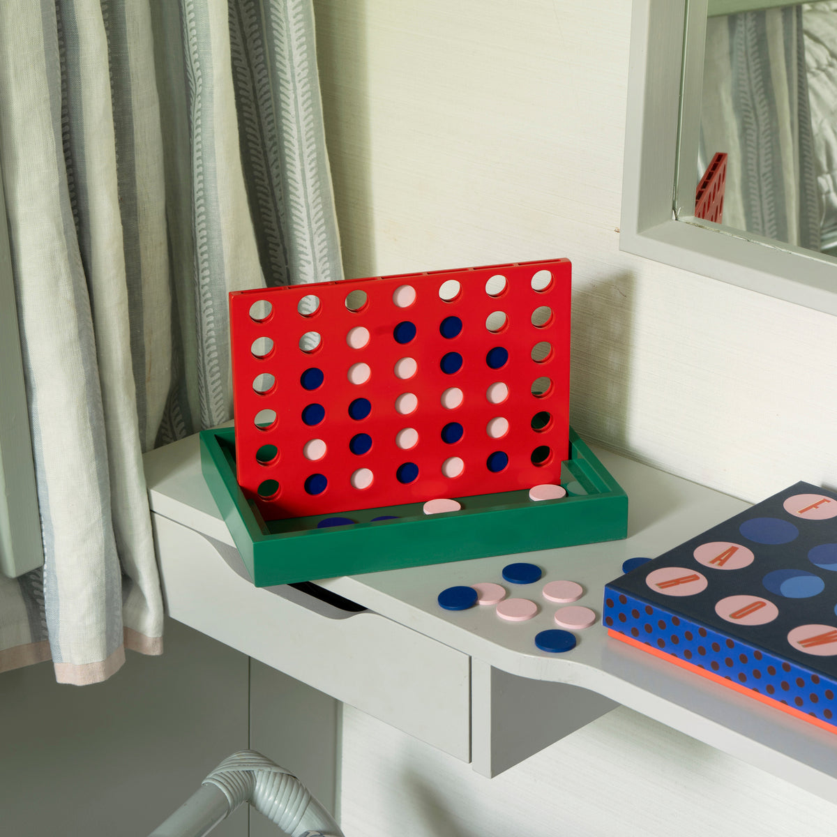 A colorful Connect 4 game set displayed on a white desk next to a window with gray curtains. The game features a red grid board, a green base tray, blue and pink discs, and a patterned storage box.