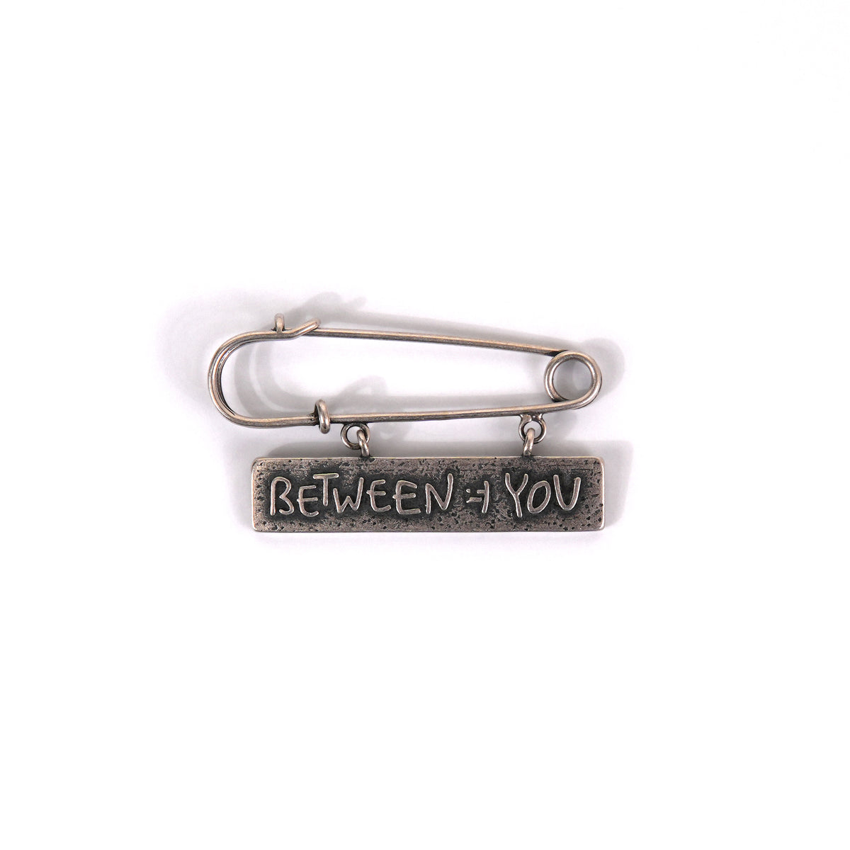 A silver-toned metal safety pin brooch with a rectangular bar engraved with the words 'BETWEEN + YOU' in raised lettering, shown on a plain white background.