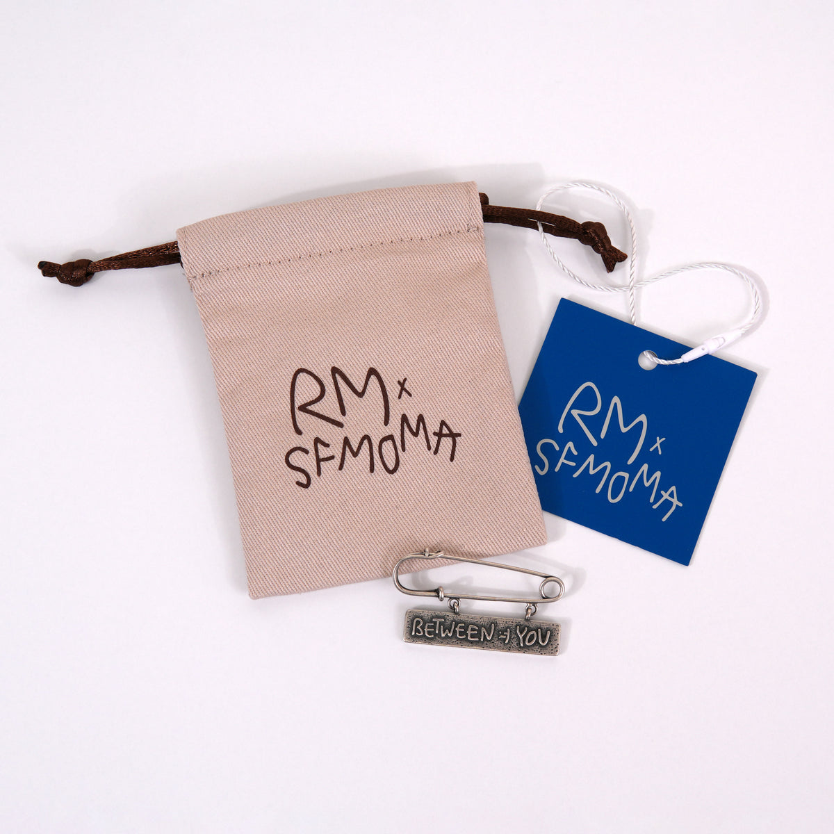 A silver metal safety pin brooch engraved with 'BETWEEN YOU' is displayed with its beige drawstring pouch, a blue paper tag, and a silver chain on a white background. The pouch and tag feature the brand name 'RM x SFMOMA'.