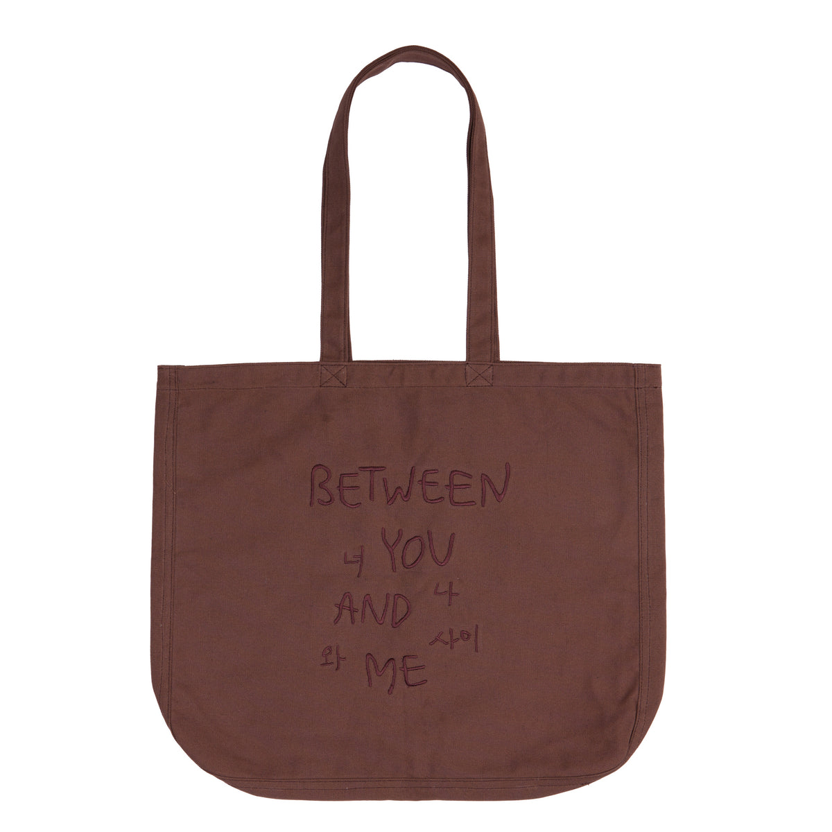 A brown canvas tote bag with two shoulder straps, featuring the text 'BETWEEN YOU AND ME' printed in a dark brown handwritten-style font on the front, set against a plain white background.