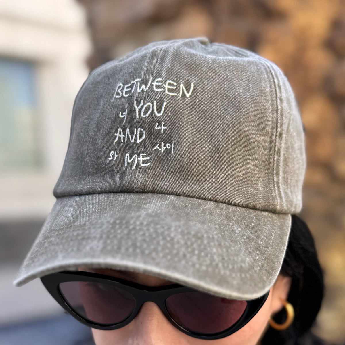 A close-up of a person wearing a gray acid-wash baseball cap with white embroidered text reading 'BETWEEN YOU AND ME' and black sunglasses against a blurred outdoor background.