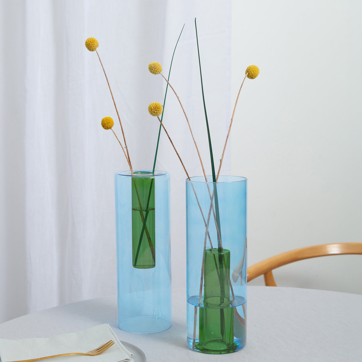Two translucent cylindrical glass vases in light blue and medium blue, each containing green stems with small yellow pom-pom flowers, set on a white table against a soft white curtain background.