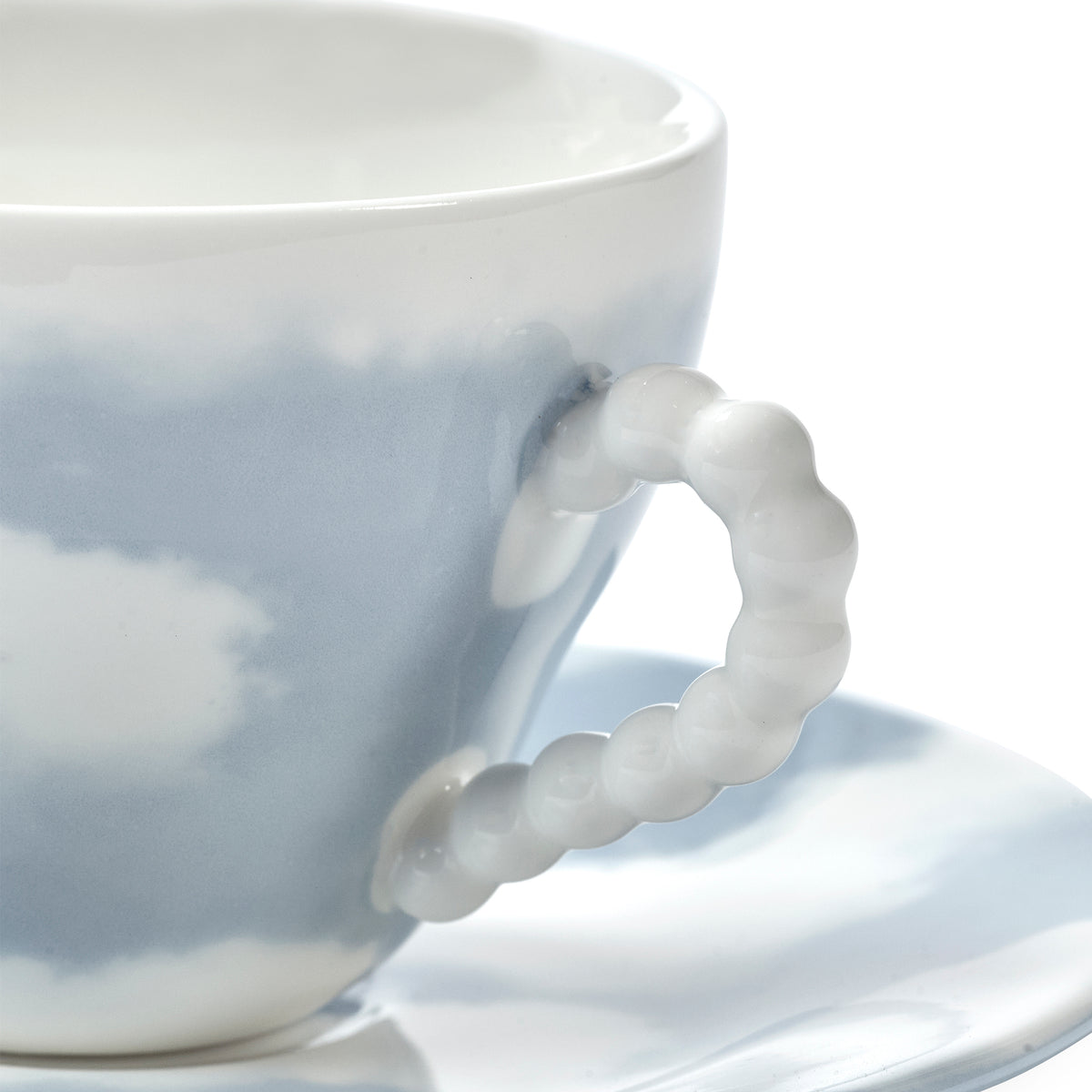 Close-up of a white ceramic teacup with a light blue cloudy pattern and a sculptural, rounded beaded handle, resting on a matching saucer against a white background.