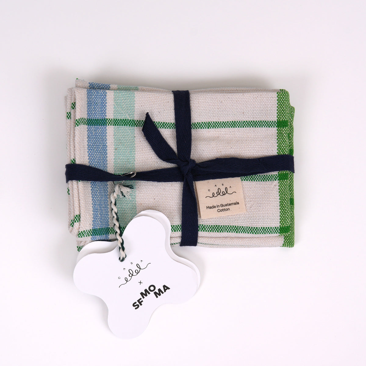 Folded placemats with a green and blue plaid pattern on a cream background, tied with a navy ribbon and a white cloud-shaped tag that reads 'Casa Edel x SFMOMA.