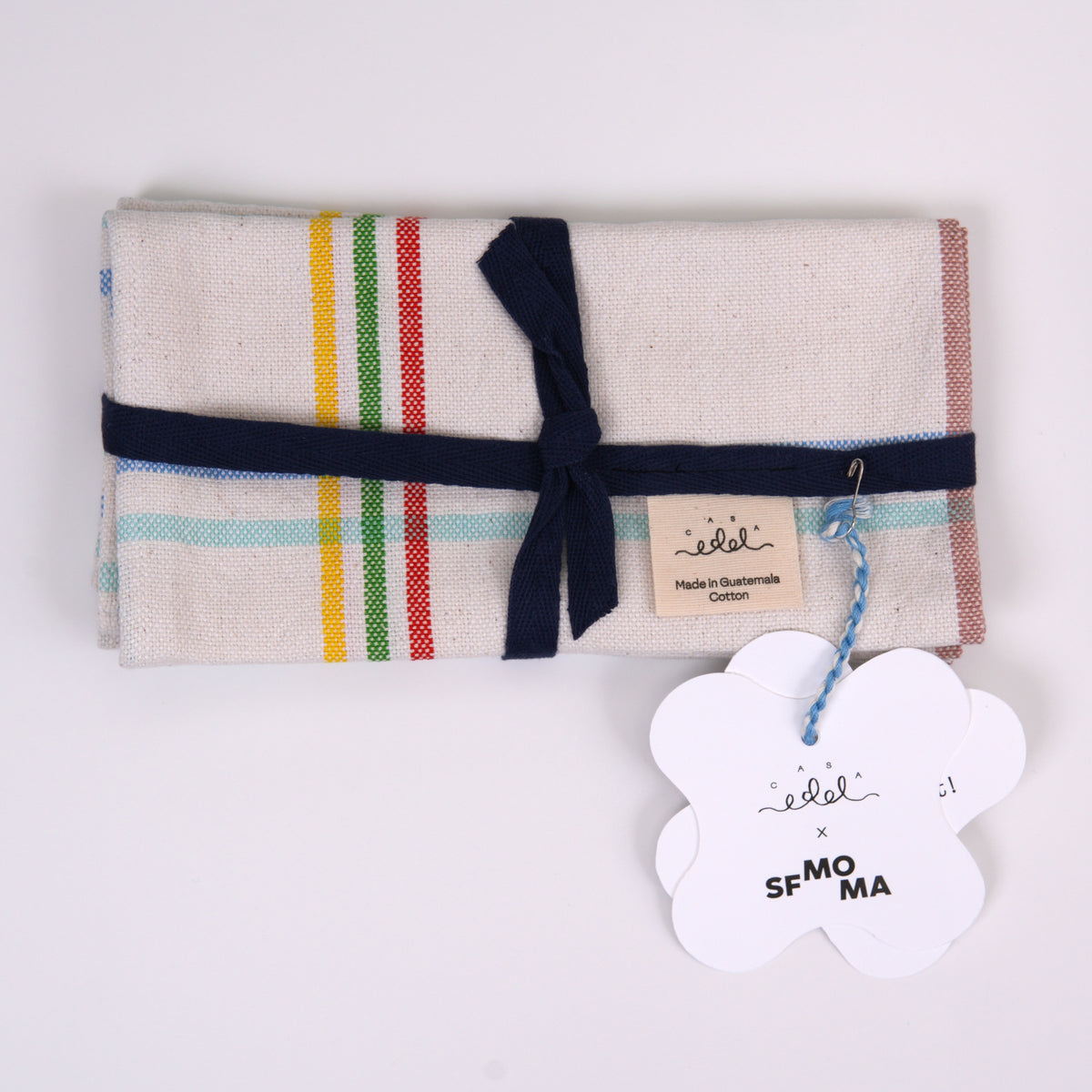A folded cotton napkinwith a cream base and multicolor stripes (green, yellow, red, light blue, and pink), tied with a navy blue ribbon and a paper tag, on a white background.
