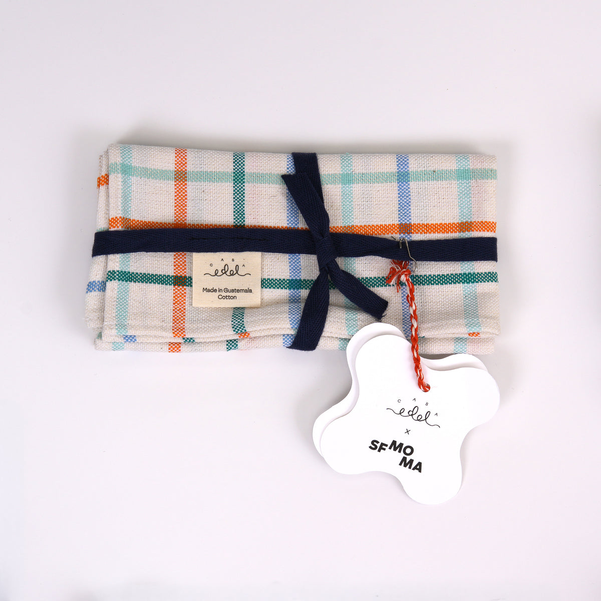 A folded cotton cloth with a multicolor plaid pattern in orange, teal, and navy on a cream base, tied with a navy ribbon and featuring a white bone-shaped paper tag.
