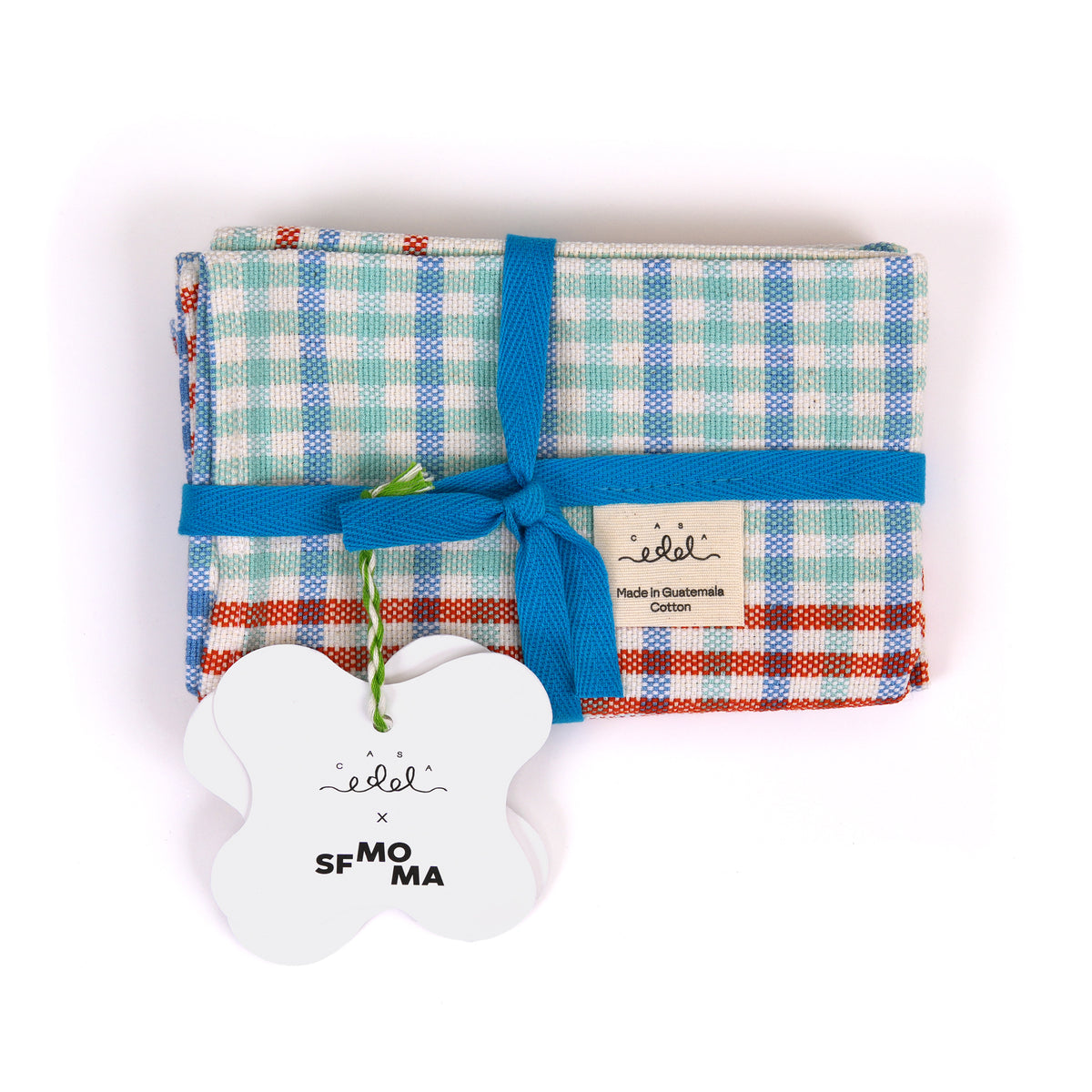 A folded placemat with a blue, green, and orange plaid pattern, wrapped in a blue ribbon with a white tag and a white bone-shaped tag that reads 'edel x SFMOMA'.