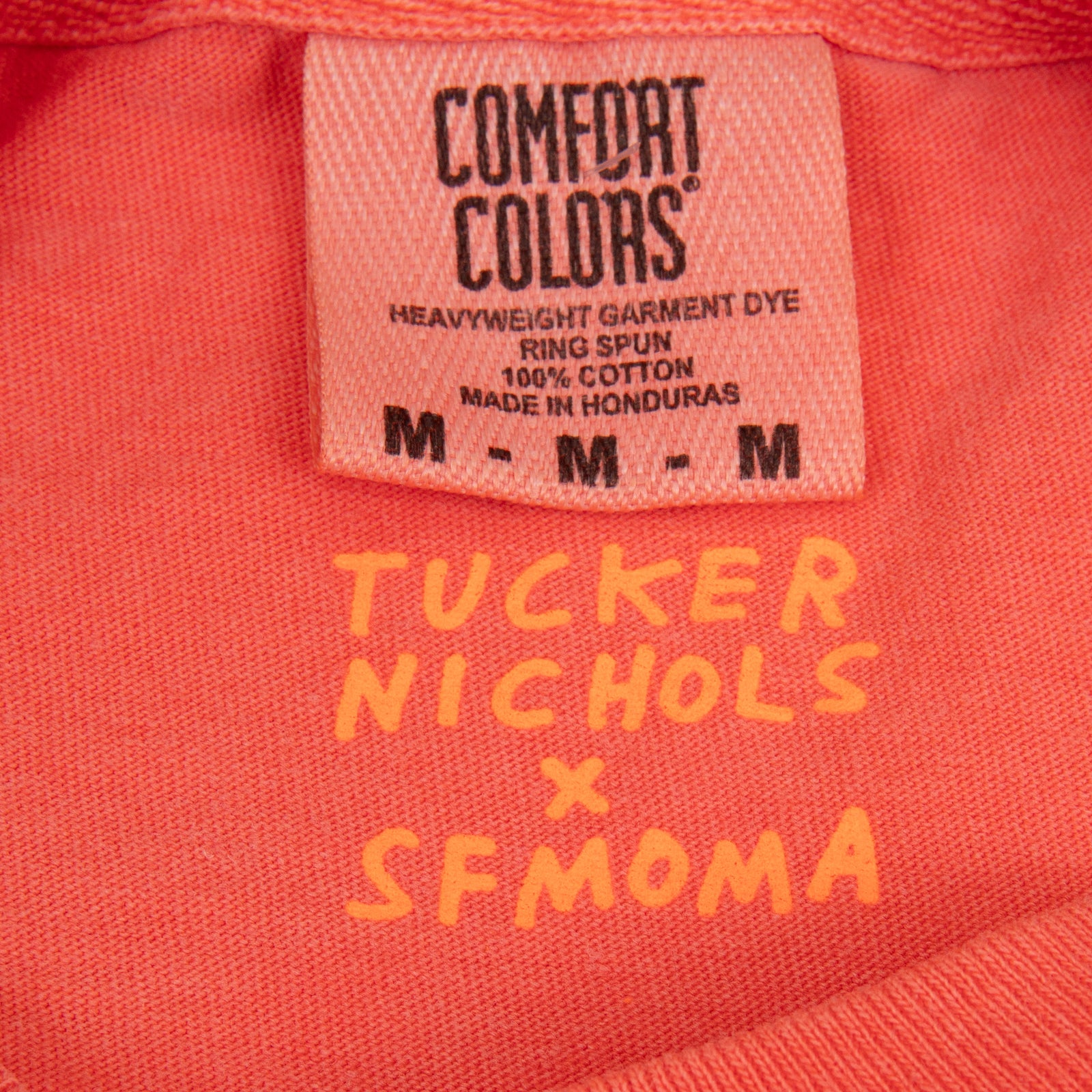 A bright coral orange crewneck t-shirt featuring the words 'San Fran' printed across the chest in a vibrant pink script font, displayed flat against a solid white background.