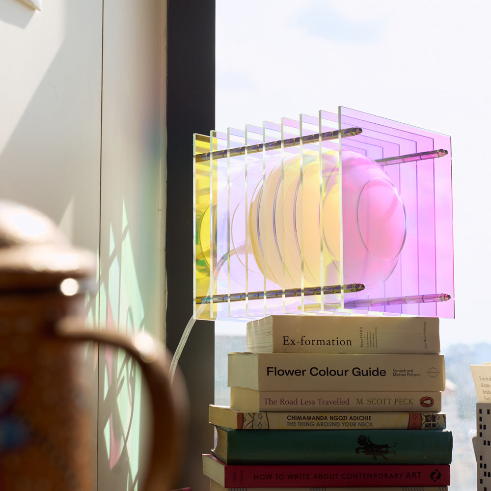 Stack of books with a colorful Iridescent lamp on top, near a window with a blurred view.