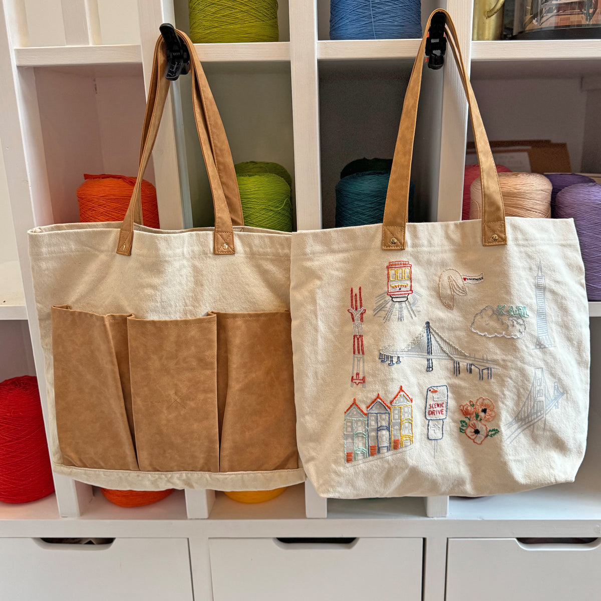 Two tote bags, one with brown pockets and the other embroidered with designs, on a shelf with colorful yarn balls.