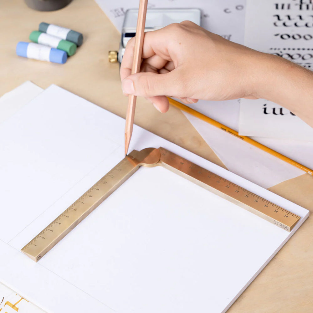 Hand holding a pencil with a brass ruler on a piece of paper, with stationery items in the background.