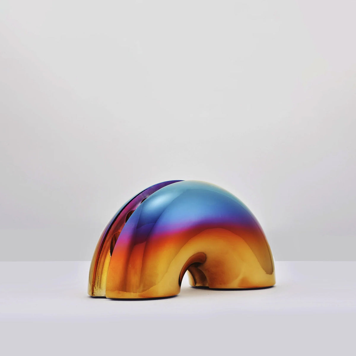 A glossy, dome-shaped menorah with a smooth iridescent finish showing a gradient of gold, orange, purple, and blue, set against a plain light gray background.