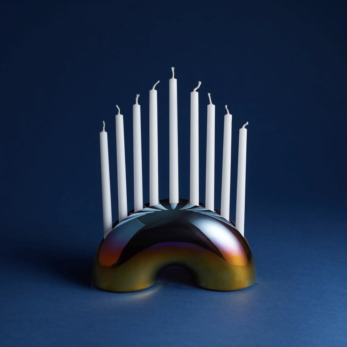 A modern arched menorah with a reflective, iridescent metallic base and nine white taper candles standing upright, set against a dark blue background.
