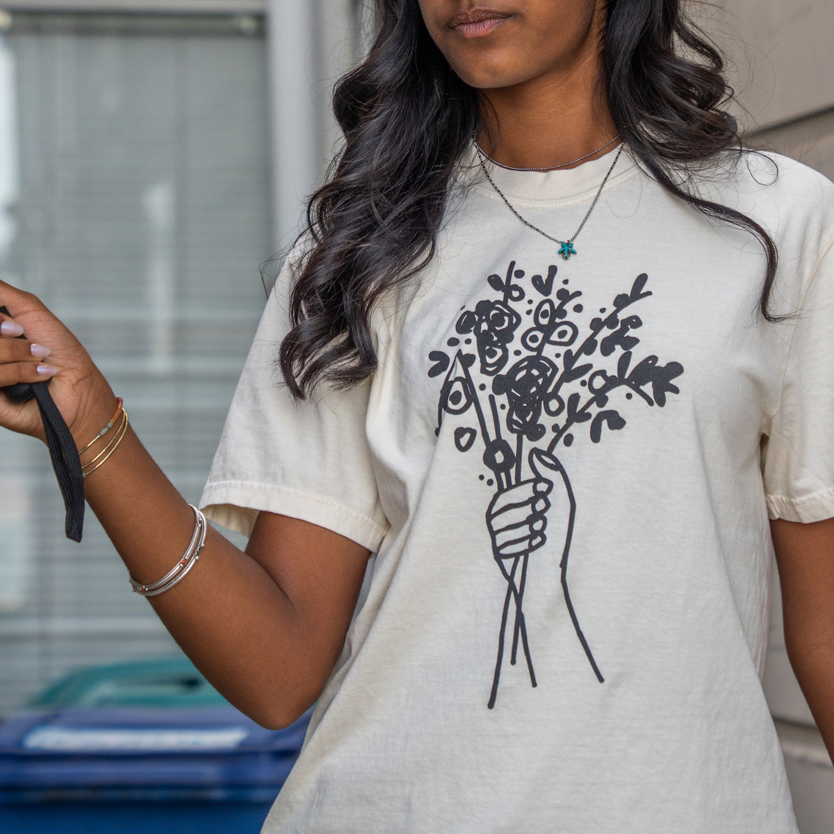 A woman wearing a cream-colored t-shirt with a black line art graphic of a hand holding a bouquet of flowers, photographed in an outdoor setting.