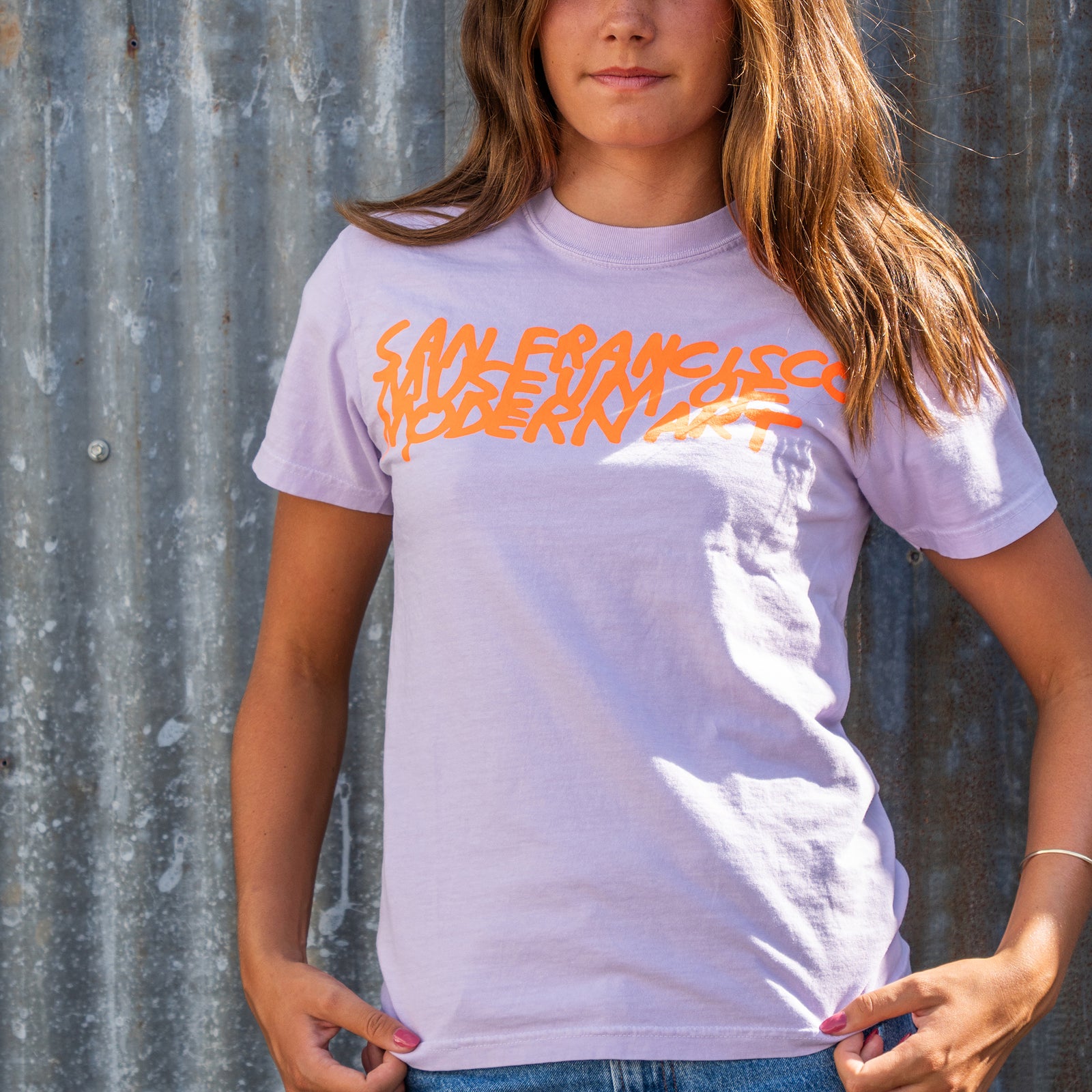 A light purple crewneck t-shirt featuring the text 'SAN FRANCISCO MODERN ART' printed across the chest in a bold, orange graffiti-style font, displayed flat against a white background.